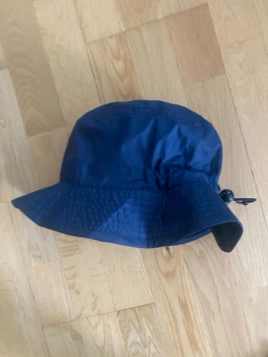 Mörkblå bucket hat med dragsko - Snygg mörkblå bucket hat i lätt och vattenavvisande material. Hatten har bred skärm och justerbar dragsko baktill för perfekt passform. Perfekt accessoar för att lyfta din stil och skydda mot sol eller regn.