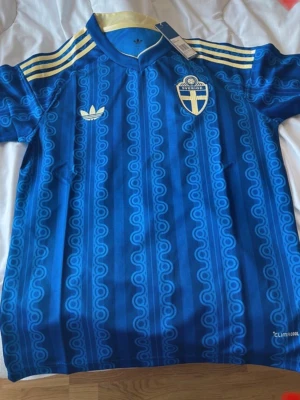 Blå Sverige fotbollströja Adidas - Säljer en blå fotbollströja från Adidas med Sveriges landslagsemblem på bröstet och gula detaljer på axlarna. Tröjan har ett retroinspirerat mönster, korta ärmar och ClimaCool-material som andas. Perfekt för match eller träning.ett till ett ,fraktar snabbt :)