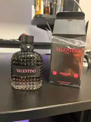 Valentino Uomo Born In Roma Intense EdP - Valentino Uomo Born In Roma Intense Eau de Parfum i en snygg svart glasflaska med nitar och matchande svart kartong. Flaskan rymmer 100 ml och har en lyxig, modern design med rosa detaljer på etiketten och förpackningen.