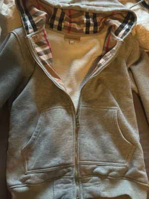 Grå bb hoodie med dragkedja och mönstrad huva - Säljer en grå hoodie från Burberry med dragkedja och fickor framtill. Insidan av huvan har det klassiska Burberry-rutiga mönstret i beige, rött, svart och vitt. Hoodien är i mjukt material och har ribbade muddar vid ärmar och nederkant.
