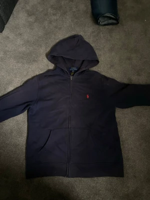 Mörkblå hoodie från Polo Ralph Lauren - Snygg mörkblå hoodie med dragkedja från Polo Ralph Lauren. Klassisk design med huva, kängurufickor och röd broderad logga på bröstet. Perfekt för en chill och stilren look.