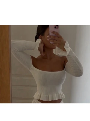 Vit ribbad topp med volang - Säljer en vit ribbad topp med långa ärmar och volangdetaljer ärmslut och nederkant. Toppen har en figurnära passform och offshoulder-design. 