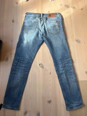 Blå slim fit jeans med slitningar - Säljer ett par blå slim fit jeans med snygga slitningar och tvättade detaljer. Jeansen har klassisk femficksmodell, normal midja och raka ben. Materialet är denim i bomull och de har en skinnpatch bak i linningen. Storleken är: W29 L30