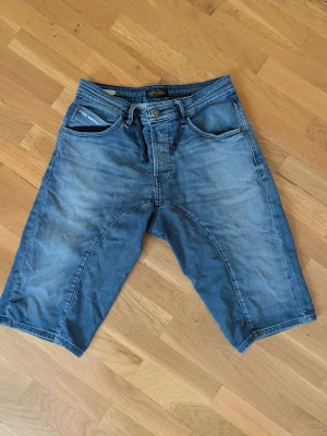 Blå jeansshorts från Jack & Jones - Säljer ett par blå jeansshorts från Jack & Jones med loose fit och snörning i midjan. Klassisk femficksmodell med knappgylf och slitningar för en avslappnad look. Perfekta för dig som gillar en bekväm och ledig stil.