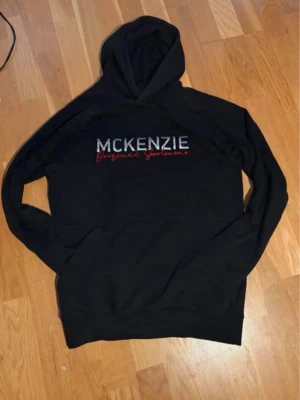 Svart hoodie från McKenzie - Svart hoodie från McKenzie med broderad logga och texten 'Original Sportswear' i vitt och rött på bröstet. Klassisk huva och mjukt material, perfekt för en avslappnad stil. Ribbade muddar vid ärmslut och nederkant.