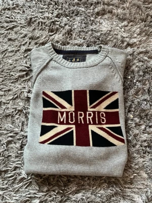 Grå stickad tröja Morris - Snygg grå stickad tröja från Morris med Union Jack-flagga och Morris-text på bröstet. Tröjan har rund hals, ribbade muddar vid ärmslut och nederkant samt detaljerade sömmar på ärmarna. Perfekt för dig som gillar klassisk men trendig stil.