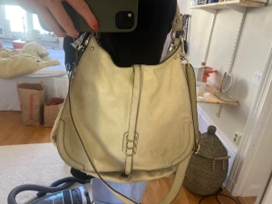 Beige skinnväska med gulddetaljer - Stilren beige handväska i skinn med både axelrem och handtag. Väskan har guldfärgade metalldetaljer och en dekorativ rem framtill. Rymlig modell med mjuk form och svarta kanter som kontrast.