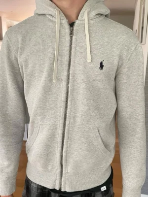 Grå zip hoodie från Polo Ralph Lauren - Snygg Ralph lauren hoodie som passar till allt. Använd under sommaren men har tröttnat på den nu. Bra skick. Passar mig som är 173 och väger 65. Storlek s