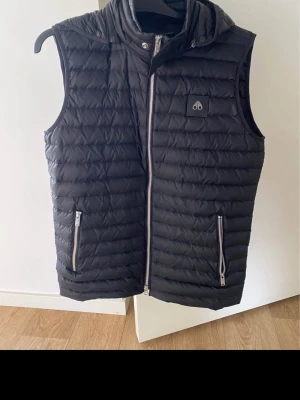 Moose knuckles Svart dunväst med huva - ÄKTA Aldrig använd Stilren svart dunväst med huva och två dragkedjeförsedda fickor framtill. Västen har quiltad design och en diskret logotyp på bröstet. Perfekt för lager-på-lager och håller dig varm under kyliga dagar.