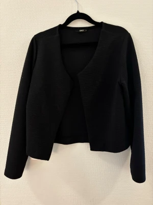 Svart öppen kofta från ONLY - Stilren svart öppen kofta från ONLY med lång ärm och enkel, rak passform. Koftan är tillverkad i ett mjukt, stretchigt material och har en minimalistisk design utan knappar eller dragkedja. Perfekt att slänga över en topp för en clean look. Fint skick. Storlek M