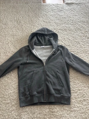 Full-zip hoodie från Mango - Mörkgrå hoodie från MNG i storlek M. Tröjan har dragkedja framtill, två stora fickor och en mysig huva med dragsko. Insidan är fodrad med mjukt, ljust tyg som gör den extra bekväm. Perfekt för chill dagar eller när du vill ha något enkelt och snyggt. Använts fåtal gånger så så gott som ny.