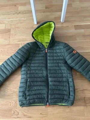 Grön pufferjacka från Save The Duck - Snygg grön pufferjacka från Save The Duck med huva och dragkedja. Insidan är limegrön och jackan har ett märke på ärmen. Tillverkad i 100% animal free material och perfekt för kalla vinterdagar. Jackan har en modern, quiltad design och två sidofickor.