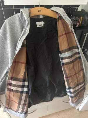 Grå hoodie med dragkedja och mönstrat foder - Snygg grå hoodie med dragkedja och huva. Insidan har ett klassiskt rutigt foder i beige, svart, vitt och rött. Utsidan är enkel och stilren med fickor framtill och dragsko i huvan. Perfekt för dig som gillar en clean look med en twist.