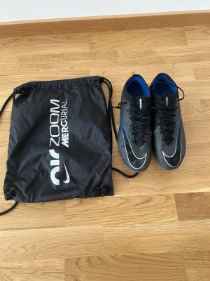 Nike Mercurial Air Zoom fotbollsskor strl 44 - Säljer ett par Nike Mercurial Air Zoom fotbollsskor i svart med blå och vita detaljer. Skorna har snörning, låg profil och är tillverkade i ett syntetmaterial för bra grepp och känsla på planen. Medföljer en svart Nike-påse med Mercurial-tryck.