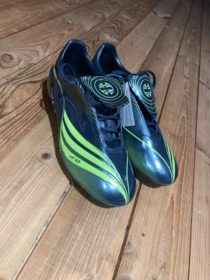 Adidas +F10 Fotbollsskor - Säljer ett par helt nya fotbollsskor i grått och limegrönt. Skorna har grå snörning och grå och limegrön sula. Skorna har en täckt plös med coolt mönster.