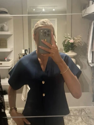Zara Mörkblå blus med guldknappar - Stilren mörkblå blus med v-ringning och två dekorativa guldknappar framtill. Blusen har korta ärmar och en rak, något boxig passform. Perfekt för en trendig och enkel look.