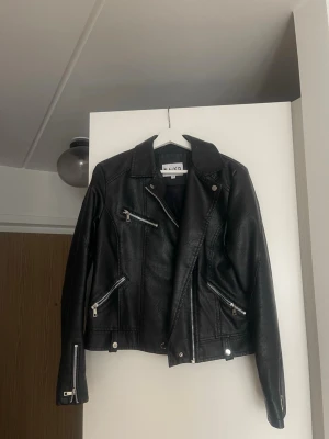 Svart skinnjacka från NA-KD - Snygg svart skinnjacka från NA-KD med flera silvriga dragkedjor och detaljer. Klassisk bikerstil med bred krage och tryckknappar. Jackan har dragkedjor vid ärmsluten och tre framfickor med dragkedja. Perfekt för en edgy look.