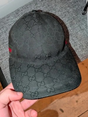 Svart Gucci keps  - Snygg svart keps från Gucci med det klassiska GG-monogrammönstret över hela kepsen. På sidan finns en grön och röd rand som ger en exklusiv touch. Justerbar rem baktill för perfekt passform. Materialet känns slitstarkt och stilrent.