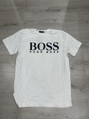 Vit BOSS t-shirt med stor logga - Vit t-shirt från Hugo Boss med stor svart BOSS-logga på bröstet. Klassisk passform och rund halsringning. Tillverkad i mjuk bomull som känns skön mot huden. Perfekt för dig som gillar stilrena och tidlösa plagg.