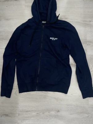 Mörkblå zip-hoodie från Replay - Snygg mörkblå hoodie från Replay med dragkedja framtill och vit logga på bröstet. Klassisk huva med svarta snören och två fickor framtill. Perfekt för en avslappnad streetwear-look. Tillverkad i mjukt bomullsmaterial.