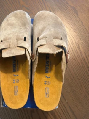 Beige Birkenstock tofflor mocka - Säljer ett par snygga beige tofflor från Birkenstock i mjuk mocka med justerbart spänne i metall. Innersulan är gul och ergonomiskt utformad för extra komfort. Perfekta för chill dagar hemma eller när du vill ha det bekvämt.