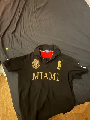 Svart pikétröja Polo Ralph Lauren - Svart pikétröja från Polo Ralph Lauren med guldfärgade broderier, stor MIAMI-text framtill och klassisk krage. Tröjan har korta ärmar och detaljer i rött på insidan av kragen. Perfekt för en sportig och stilren look.