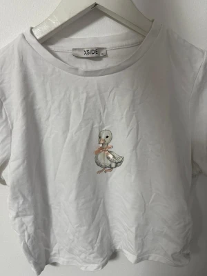 Vit croppad t-shirt med anka - Vit croppad t-shirt från XSIDE med ett gulligt tryck av en liten anka på bröstet. Klassisk rund halsringning och korta ärmar. Perfekt för en avslappnad och söt stil. Aldrig använd!!