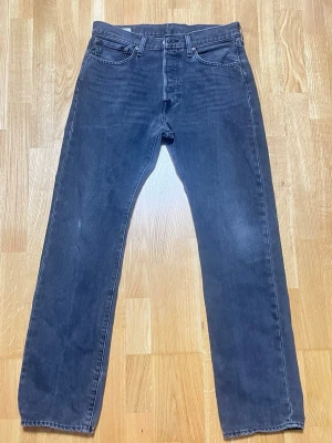 Mörk gråa/svarta Levis 501 - Säljer ett par mörk gråa/svarta jeans från Levi's. Jeansen har klassisk femficksdesign, bälteshällor och knappgylf. Materialet är slitstark denim i bomull och färgen är tvättad mörk grå för en cool look.