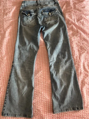 Ljusblå bootcut jeans med fickdetaljer - Säljer ett par ljusblå bootcut jeans med snygga fickor bak som har lock och dekorativa sömmar samt silverfärgade knappar. Jeansen har bälteshällor och klassisk femficksdesign. Perfekta för en avslappnad och trendig look.