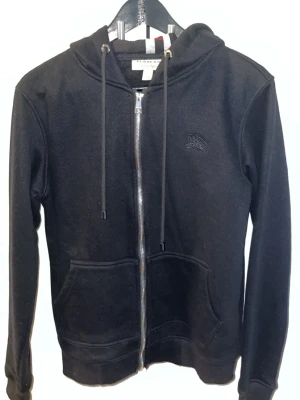 Burberry, svart hoodie med dragkedja och huva - Snygg svart hoodie med dragkedja framtill och två stora fickor. Huvan har ett klassiskt rutigt foder i svart, vitt och rött. Justerbara snören i huvan och diskret broderad logga på bröstet. Perfekt för en chill och stilren look. Storlek S men sitter lite tight.