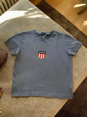 💙 Blå GANT t-shirt med tryck 💙 NYSKICK! 💙 - Snygg blå t-shirt från GANT med klassiskt tryck framtill. T-shirten har rund hals, korta ärmar och ett sköldmärke med stjärnor och ränder på bröstet. Perfekt till jeans eller shorts för en avslappnad look.