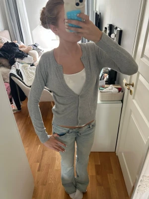 Ljusgrå basic kofta med knappar - En ljusgrå, tunn och enkel kofta med långa ärmar och knäppning framtill. Modellen är rak och har ribbade muddar vid ärmslut och nederkant. Perfekt att slänga över ett linne eller t-shirt för en chill look.