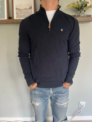Morris Stockholm Half-Zip Tröja Merinoull Marinblå S - Morris Stockholm tröja i 100% merinoull, marinblå med halv blixtlås-krage och broderad liljemärke på bröstet. Klassisk skandinavisk kvalitetsdetalj, ribbstickade kanter. OBS: mycket litet, knappt synligt hål (se bild för närbild) — påverkar inte helhetsintrycket men flaggas här för transparens.  Skick: bra skick, ett mycket litet och knappt synligt hål (se bild), i övrigt fint Material: 100% merinoull Storlek: S Modell: 175 cm / 65 kg — sitter true to size på bild