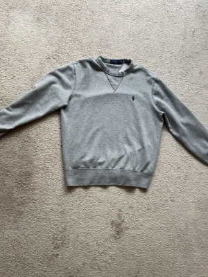 Grå sweatshirt från Polo Ralph Lauren - Klassisk grå sweatshirt från Polo Ralph Lauren med rund halsringning och blå broderad logga på bröstet. Tröjan har ribbade muddar vid ärmslut och nederkant samt en snygg detalj vid halsen. Perfekt för en avslappnad och stilren look. Säljs då den ej kommer till användning längre. Storlek M men passar även S. 