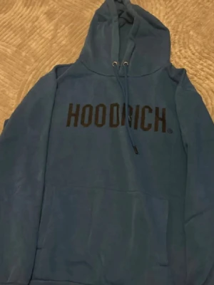 Blå HOODRICH hoodie med huva - Snygg blå hoodie från HOODRICH med stor svart logga på bröstet. Klassisk modell med huva, dragsnören och magficka framtill. Perfekt för en chill och avslappnad stil. Tillverkad i mjukt material för extra komfort.