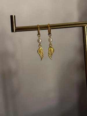 Guldiga örhängen med pärlor och vinge - Säljer ett par snygga örhängen i guldfärg med små vita pärlor och hängande vingeformade detaljer. Örhängena har en elegant och trendig look, perfekta för att lyfta vilken outfit som helst. Materialet ser ut att vara metall och pärlor.