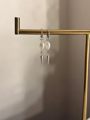 Silvriga örhängen med pärlor och spikes - Säljer ett par stilrena örhängen i silverfärg med vita pärlor och spetsiga spikes som hänger ned. Örhängena har en modern design med en cool kontrast mellan de mjuka pärlorna och de vassa detaljerna. Perfekta för dig som vill ha något unikt och trendigt.