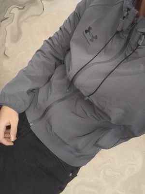Grå vindjacka från Under Armour - Snygg grå vindjacka från Under Armour med dragkedja framtill, justerbar huva och två sidofickor. Jackan har en sportig look och är tillverkad i ett lätt material som passar perfekt för blåsiga dagar. Logga på bröstet för extra stilpoäng.