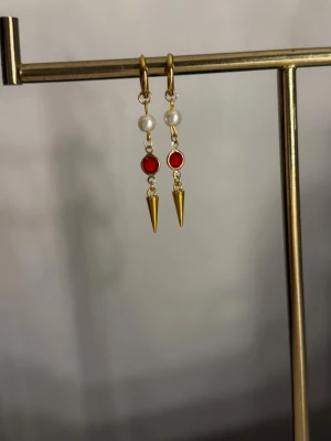 Guldörhängen med pärlor och röda detaljer - Säljer ett par unika örhängen i guldton med vita pärlor, röda runda detaljer och spetsiga hängen. Örhängena har en cool mix av klassiskt och edgy stil, perfekta för att lyfta vilken outfit som helst. Materialet ser ut att vara metall med pärlimitation och färgade stenar.