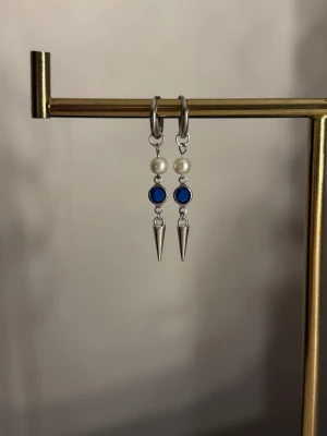 Silvriga örhängen med pärlor och blå detalj - Säljer ett par snygga silvriga örhängen med vita pärlor, blå stenliknande detalj och spetsiga hängen. Örhängena har en modern design med coola kontraster mellan metall, pärla och färg. Perfekta för dig som vill sticka ut lite extra.