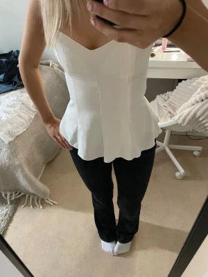 Vit peplum topp med v-ringning - Säljer en stilren vit peplum topp med v-ringning och breda axelband. Toppen har en figurnära passform upptill och faller ut nedtill för en snygg siluett. Perfekt att matcha med jeans eller kjol för en trendig look. har en liten missfärgning på kanten på baksidan men syns knappt!💕💕 