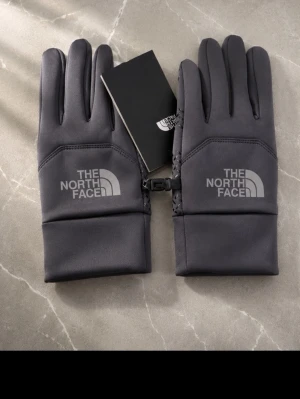 Svarta handskar från The North Face - Snygga svarta handskar från The North Face med logga på ovansidan. Handskarna har en slimmad design och är tillverkade i ett mjukt syntetmaterial. Perfekta för kyliga dagar och har ett smidigt spänne som håller ihop dem när de inte används.