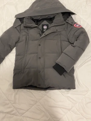 Grå dunjacka från Canada Goose - Säljer en grå dunjacka från Canada Goose med huva och klassisk logga på ärmen. Jackan har raka linjer, stora fickor framtill och stängs med både dragkedja och knappar. Perfekt för kalla vinterdagar och riktigt varm.