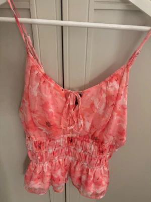Blommig rosa linnetopp med knyt - Supersöt linnetopp i rosa med blommigt mönster och tunna axelband. Toppen har smockad midja, volangkant och knyt fram. Perfekt för varma dagar och ger en romantisk vibe till din outfit.