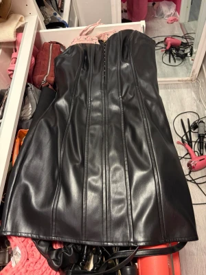 Svart axelbandslös klänning i skinnimitation - Snygg svart axelbandslös klänning i skinnimitation med markerade sömmar framtill och dragkedja. Kort modell som ger en edgy look, perfekt för dig som vill sticka ut. Klänningen har en kroppsnära passform och är tillverkad i syntetmaterial.