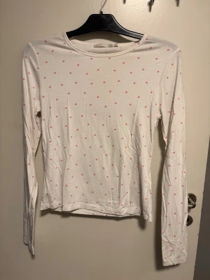 Vit långärmad pyjamas tröja  med rosa hjärtan - Söt vit långärmad pyjamas tröja  med små rosa hjärtan över hela tyget. Tillverkad i mjuk bomull och har en klassisk rund halsringning. Perfekt för dig som gillar gulliga detaljer och enkel stil.