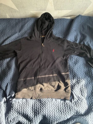 Svart hoodie från Polo Ralph Lauren - Snygg svart hoodie från Polo Ralph Lauren med dragkedja framtill och klassisk röd logga på bröstet. Hoodien har huva och känguruficka, perfekt för en snygg och stilren look. Tillverkad i mjukt material som känns skönt mot huden.