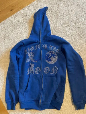 Blå hoodie med dragkedja och tryck - Säljer en blå hoodie med huva och dragkedja framtill. På framsidan finns ett stort tryck med texten 'AIM FOR THE MOON' och motiv av en tecknad figur och en måne i glittrande stenar. Hoodien har kängurufickor och ribbade muddar. Perfekt för dig som gillar streetwear och unika detaljer.