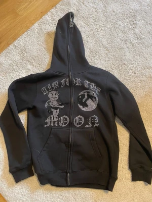 Svart hoodie med strasstryck och dragkedja - Säljer en svart hoodie med dragkedja och huva. På ryggen finns ett stort strasstryck med texten 'AIM FOR THE MOON' och en tecknad figur med vingar och krona samt en måne. Hoodien har två fickor fram och ribbade muddar. Perfekt för dig som gillar streetwear och unika detaljer.