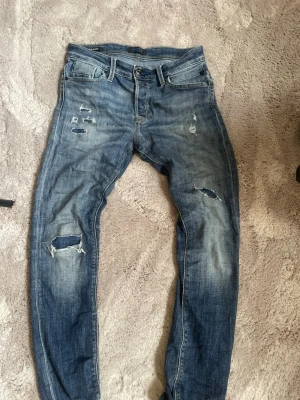 Blå slitna jeans med hål - Säljer ett par blå replay jeans med slitningar och hål på både fram- och baksida. Jeansen har en klassisk femficksmodell, normal midja och raka ben. Materialet är denim i bomull 